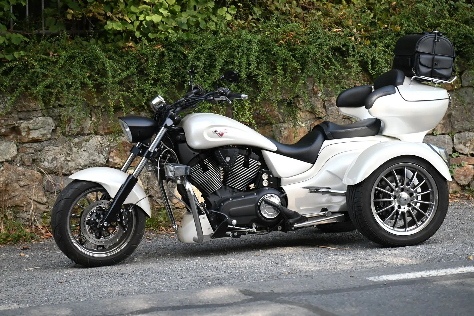 Un trike bianco a tre ruote col bauletto, parcheggiato davanti a un muro di pietra